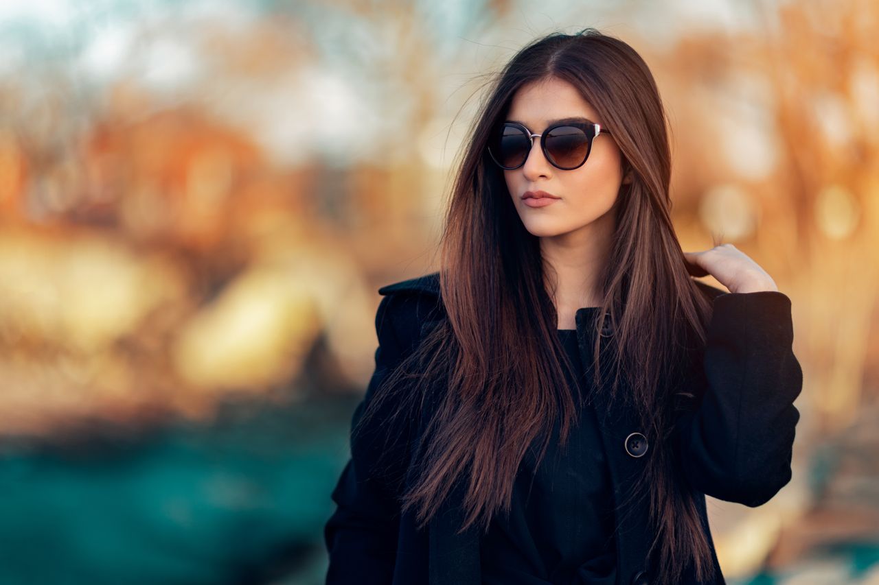 Fashionable woman wearing sunglasses and black coat, posing outdoors in natural light.