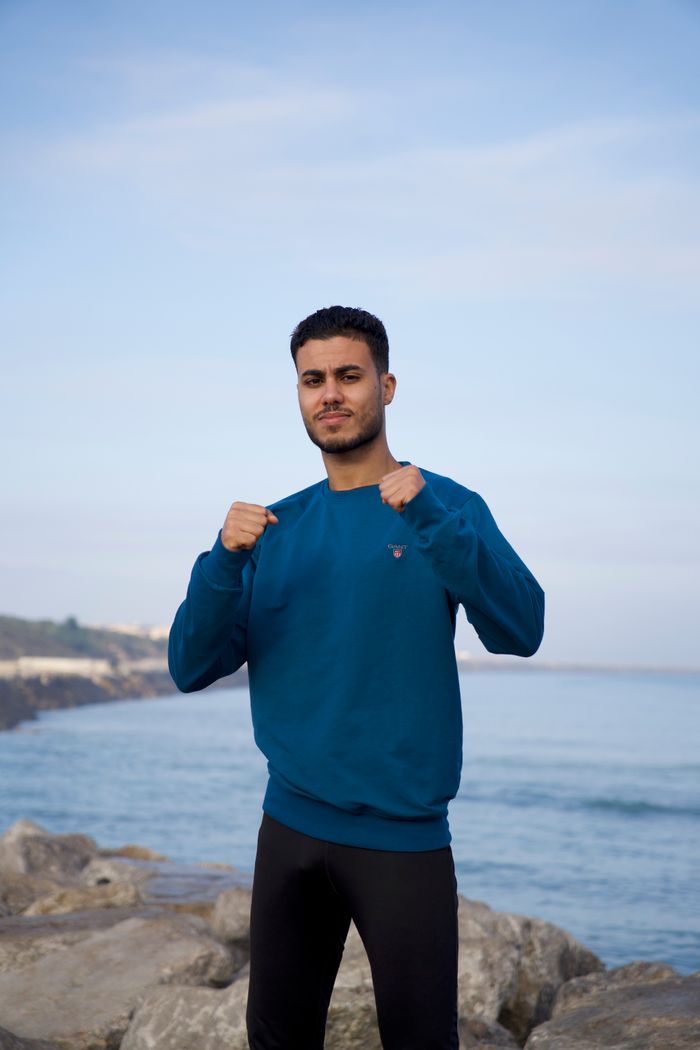 Confident young man poses on rocky ocean shore wearing a blue sweater and workout gear.