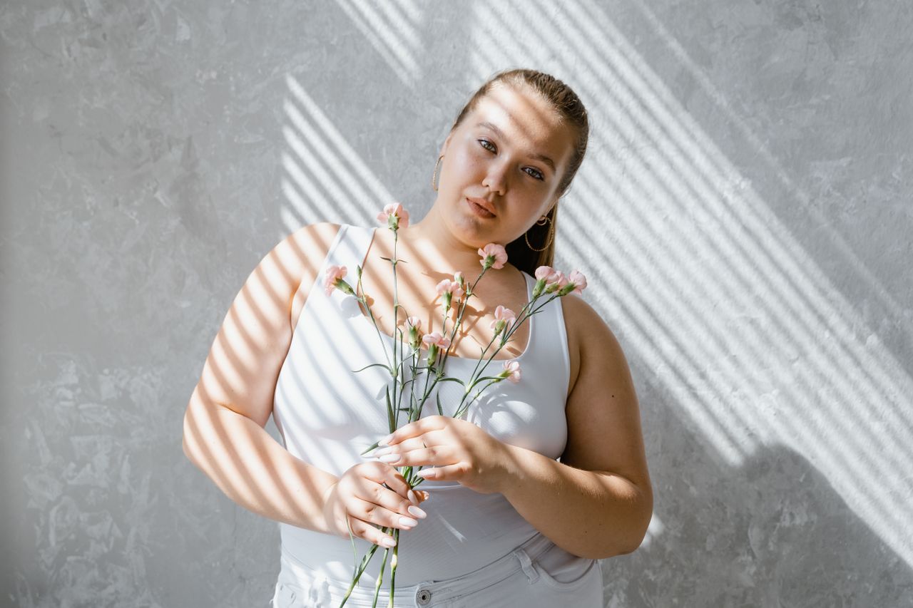 Plus-size woman in tank top holding flowers with sunlight filtering through blinds.